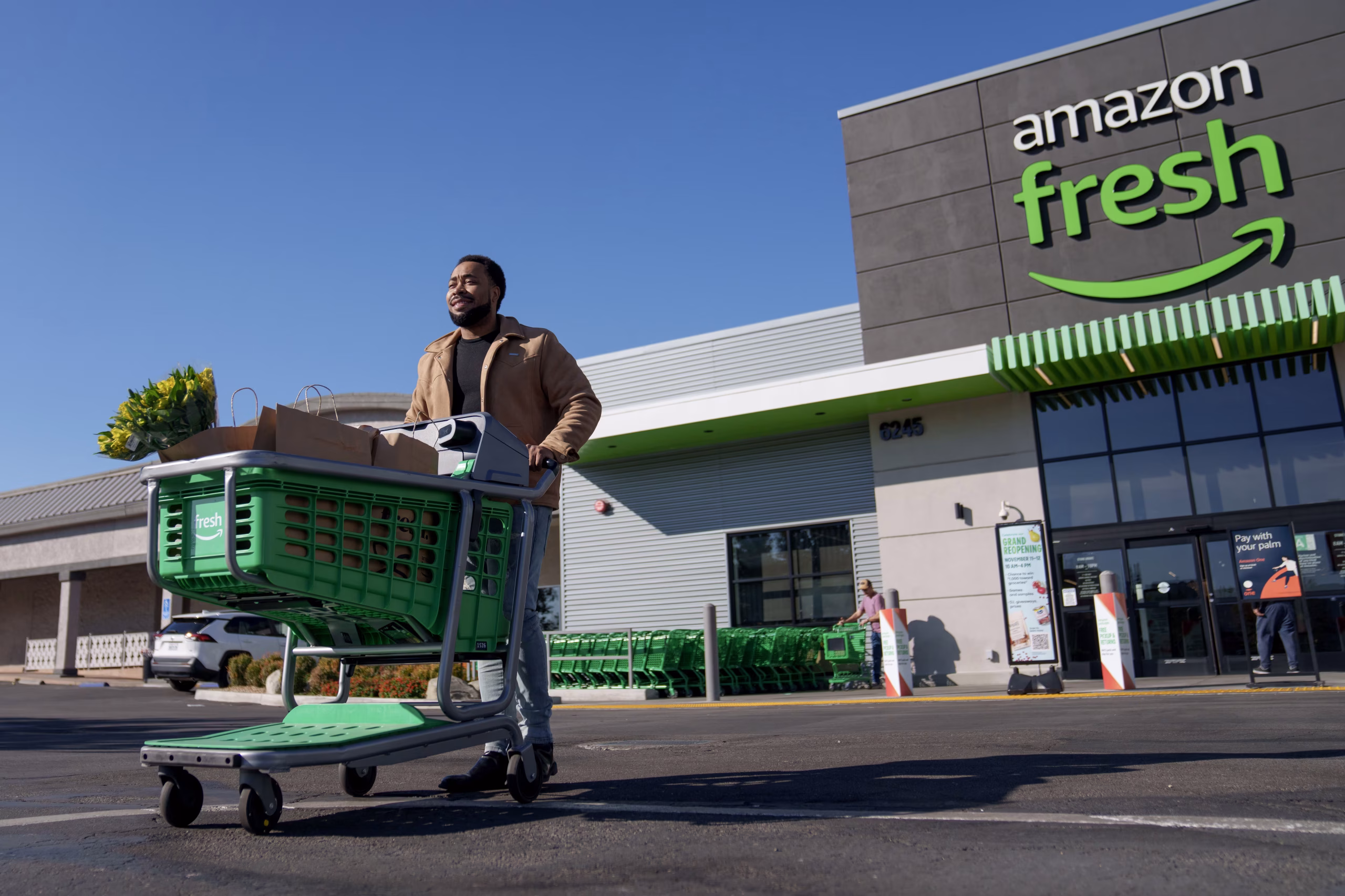 Une photo en contre-plongée montrant un homme poussant un chariot de courses vert rempli de sacs en papier brun et d'un bouquet de fleurs jaunes. Il quitte un magasin Amazon Fresh moderne au design gris et vert sous un ciel bleu clair. En arrière-plan, on aperçoit d'autres chariots alignés et l'entrée du magasin avec des affiches promotionnelles.